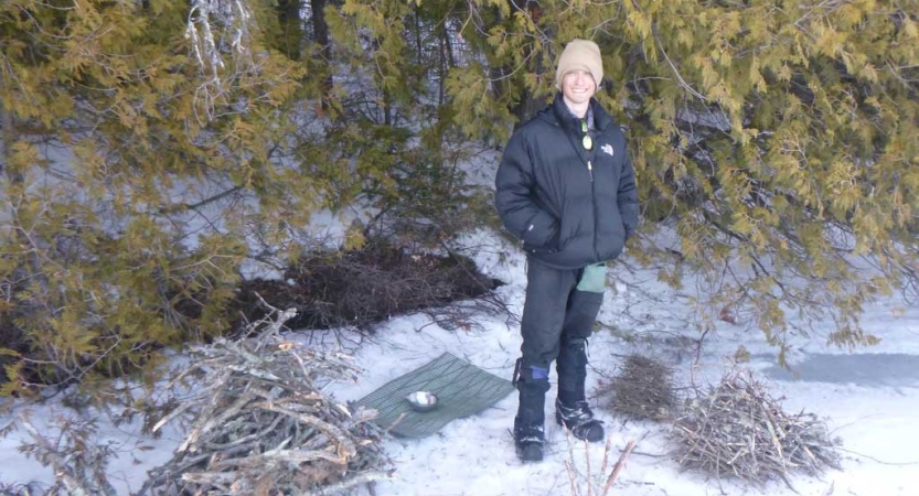 A person stands in a wooded, snowy area beside small piles of wood and a sleeping pad. 